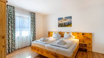 Ein Bett aus Holz mit weißen Leinen und Handtüchern, ein Nachttisch, ein Gemälde mit Berglandschaft und leichte Vorhänge mit Blätermotiv.