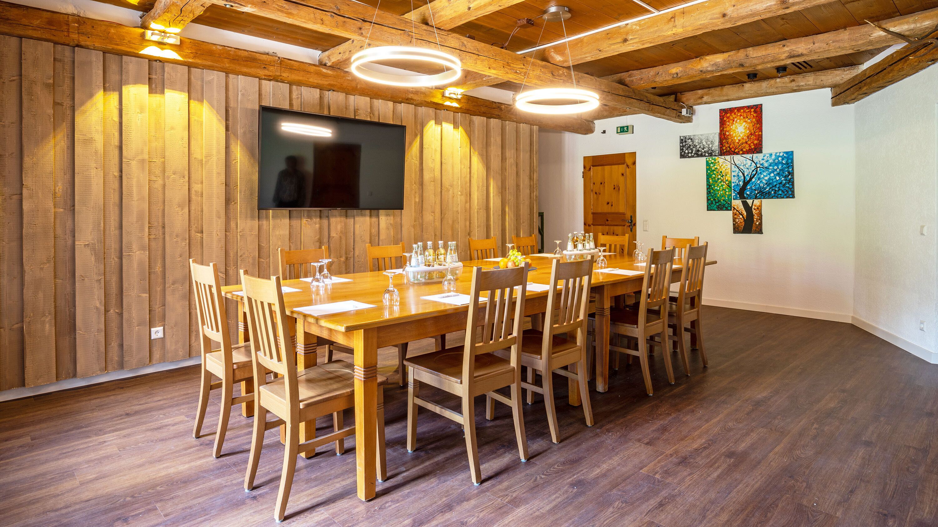 A conference room with a long wooden table, chairs, a flat-screen TV, wooden beams, and artwork on the walls.