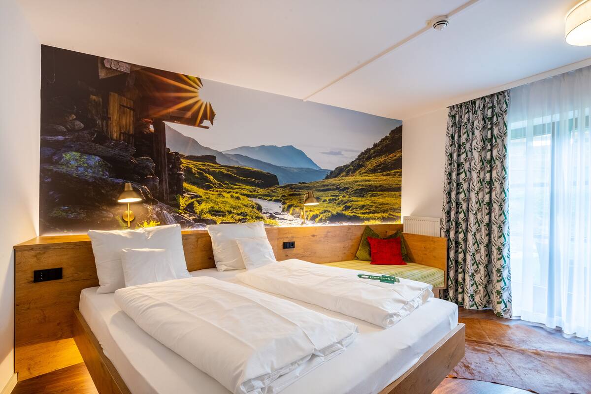 Une chambre avec un grand lit, une tête de lit en bois, deux tables de chevet avec des lampes, un oreiller rouge et un grand tableau représentant un paysage accroché au mur.