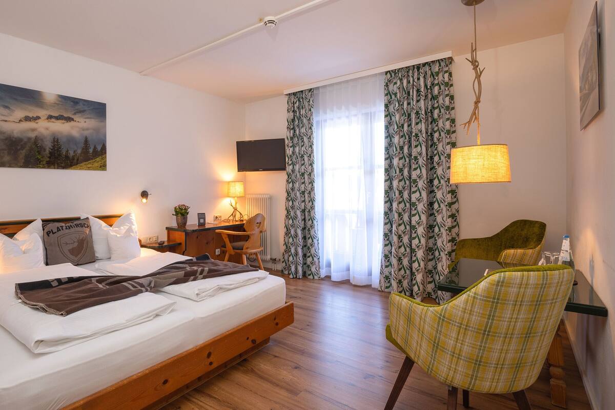 Une chambre bien éclairée, avec un lit, un bureau, une chaise et une grande fenêtre avec des rideaux.
