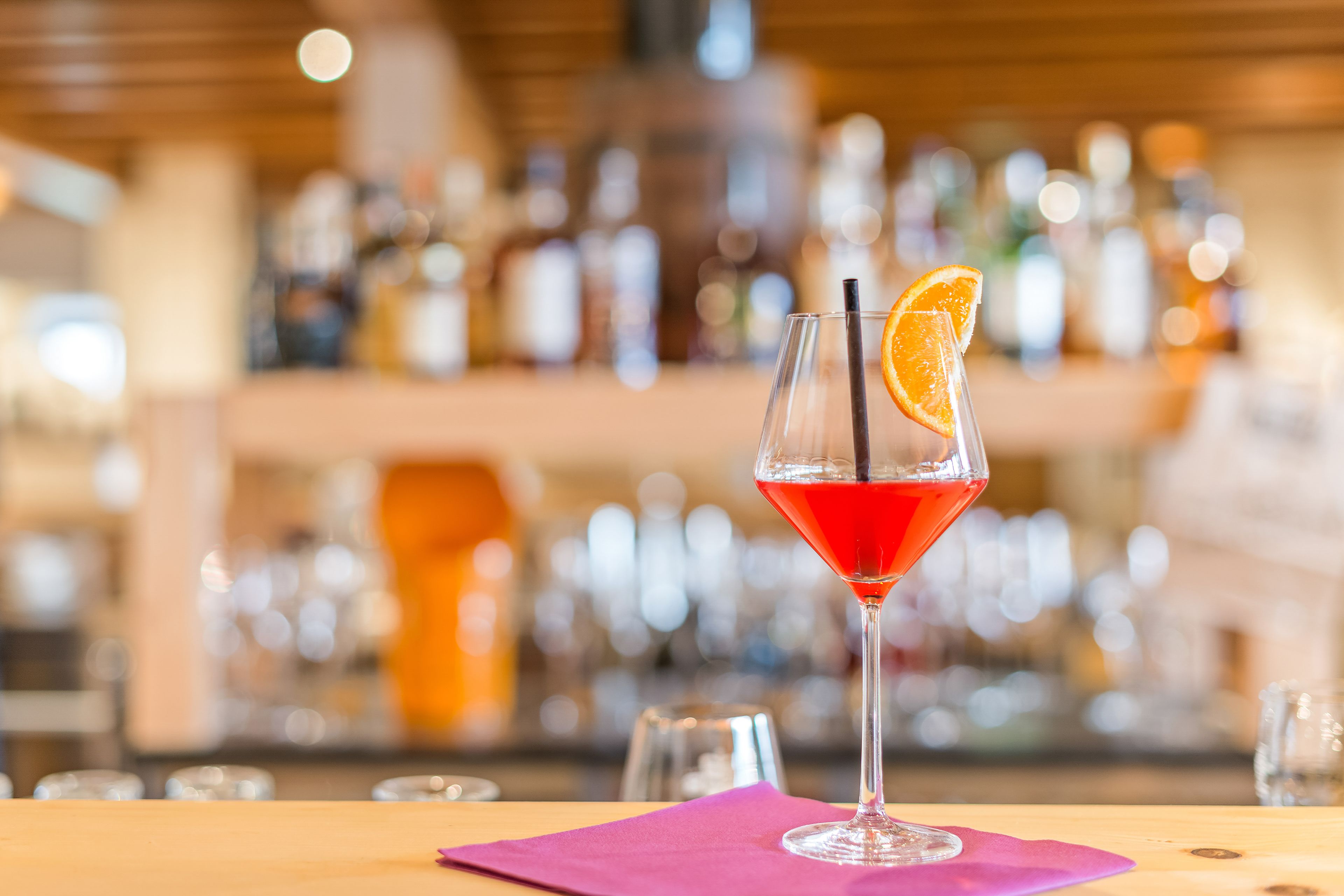 A bar setting with a cocktail glass on a wooden table, a purple napkin, and a blurred background of bottles and glasses.
