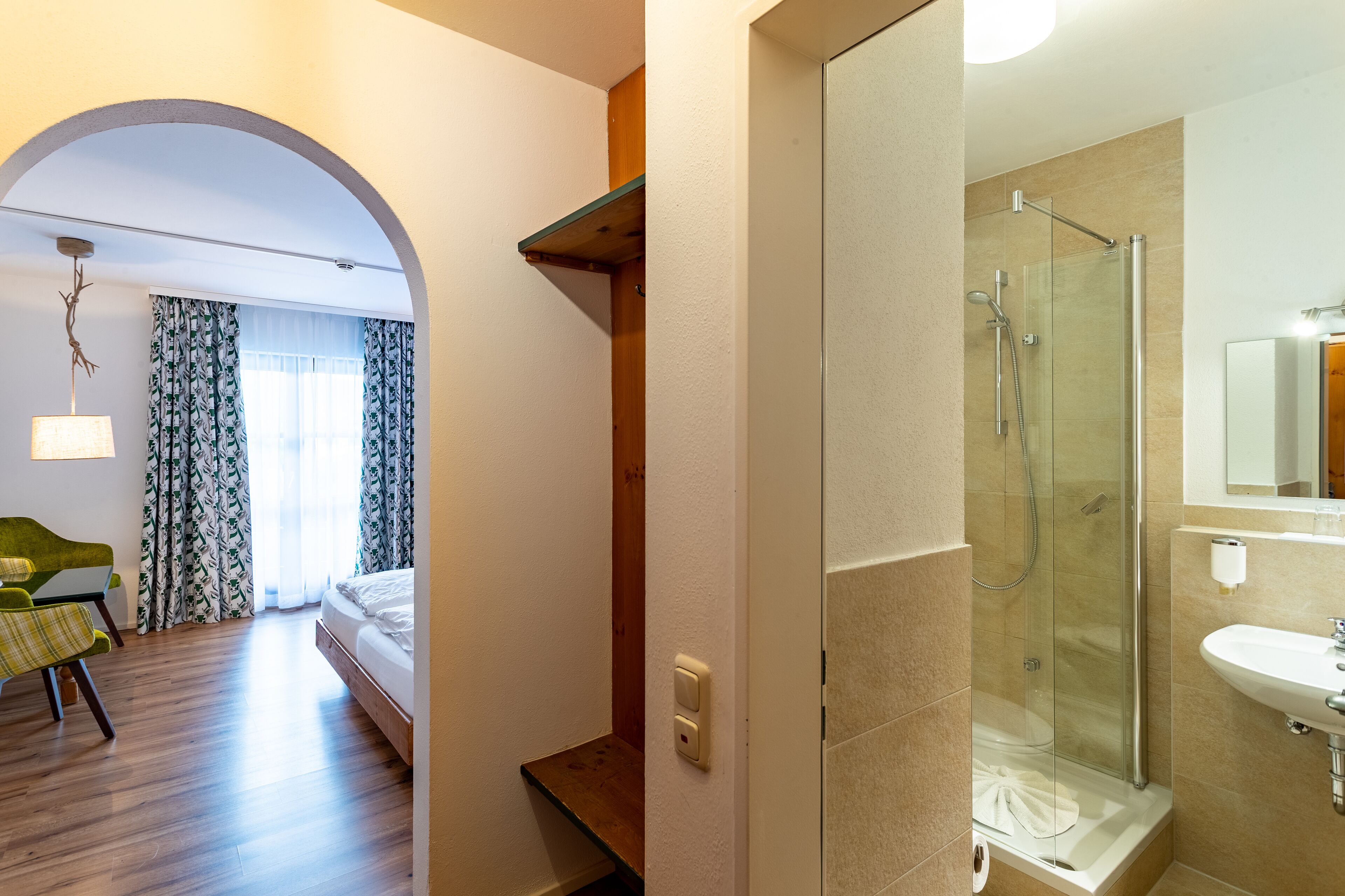 Habitación de hotel con cama, escritorio, silla y baño con ducha de vidrio y lavamanos.