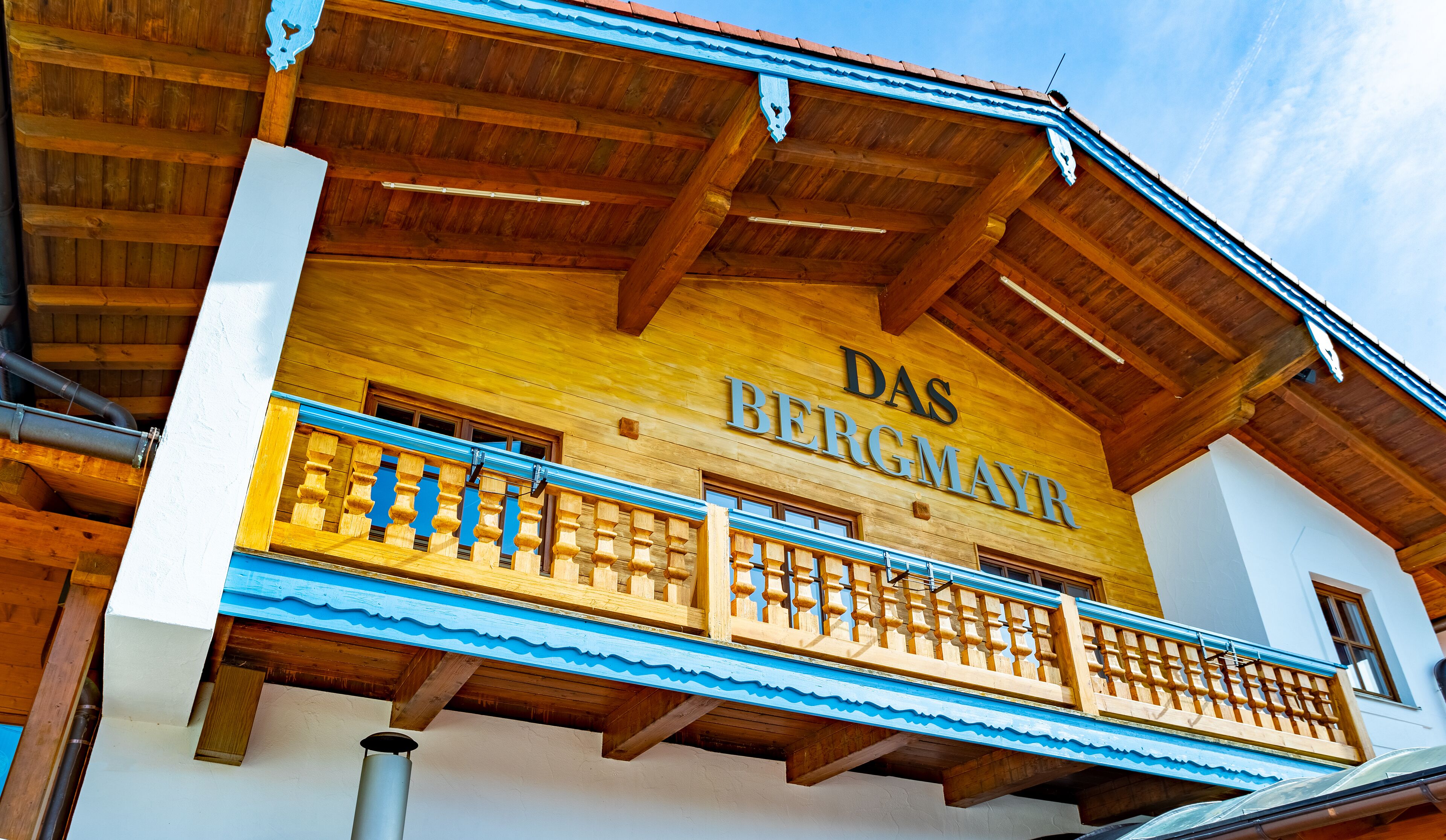 A building with a wooden facade and a balcony with a blue railing. The text "DAS BERGMAYR" is visible on the building.
