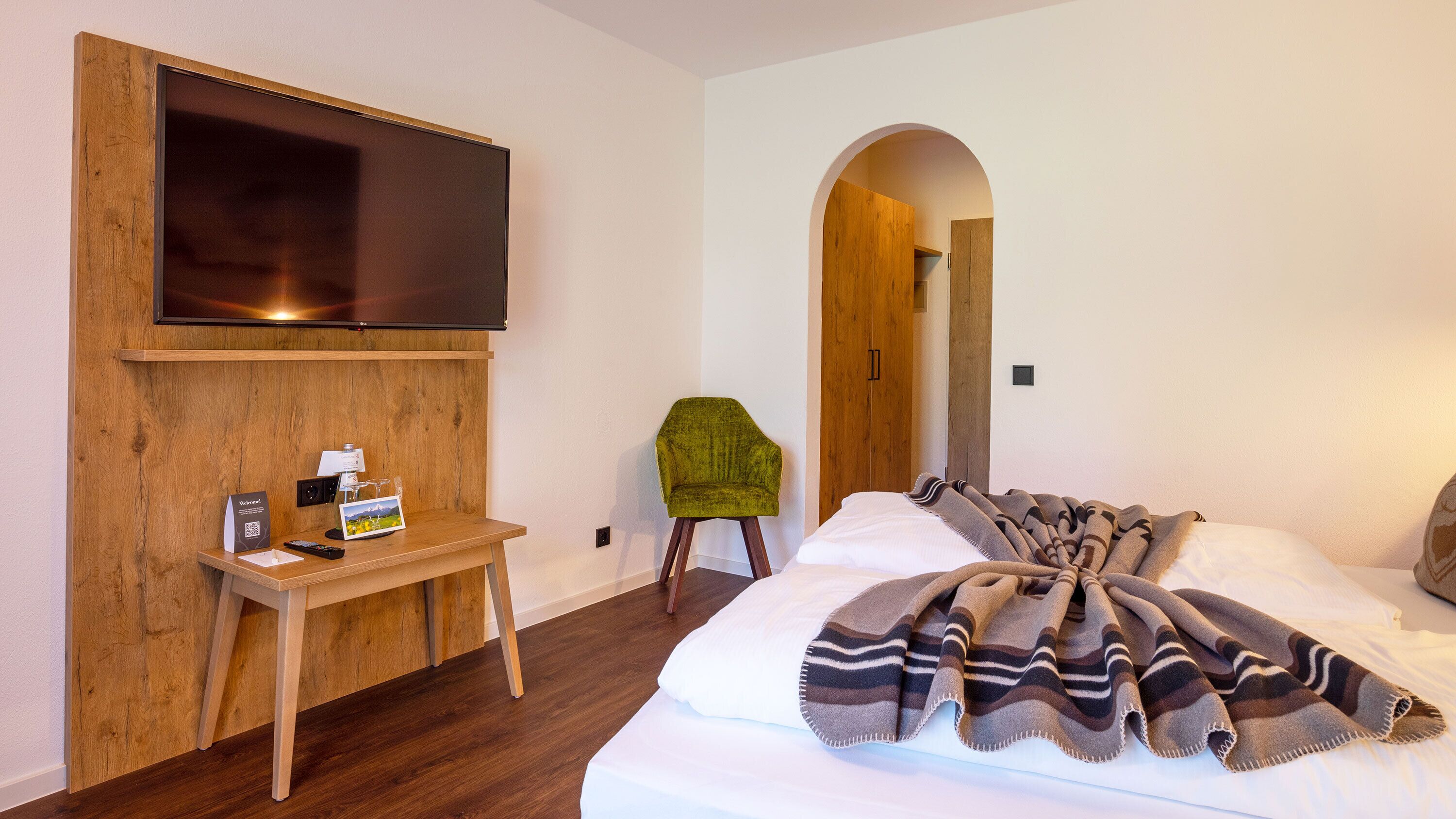 Camera d'albergo con una grande TV a schermo piatto fissata a una parete in legno, una scrivania in legno con un telecomando, una sedia verde e un letto con coperta a righe.