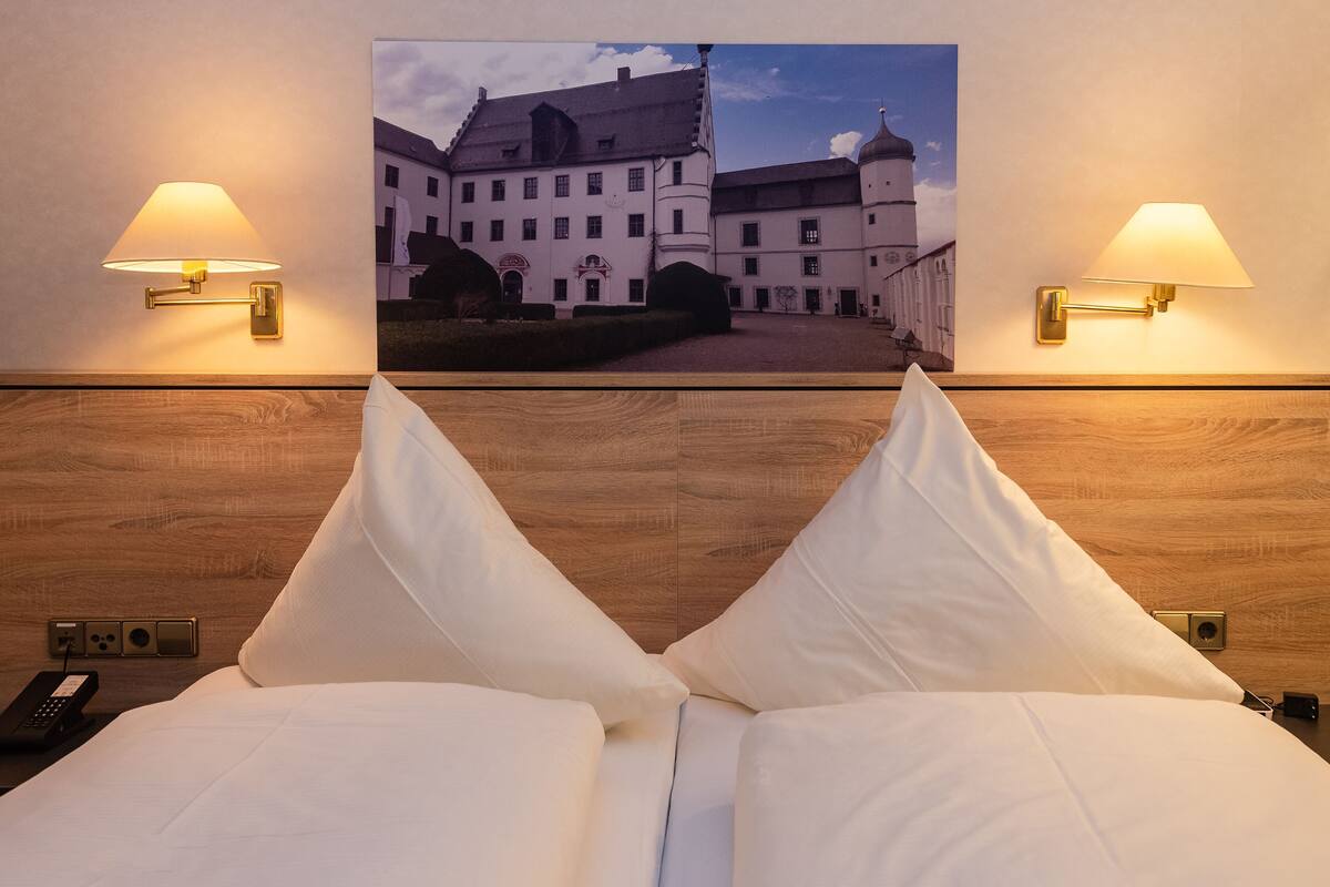 Een hotelkamer met een bed, twee wandlampen, een schilderij van een gebouw en een houten hoofdeinde.