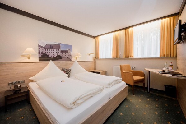 Ein Hotelzimmer mit einem Bett, zwei Nachttischen, einem Schreibtisch, einem Sessel, einem Fenster mit Vorhängen und einem an der Wand befestigten Fernseher.