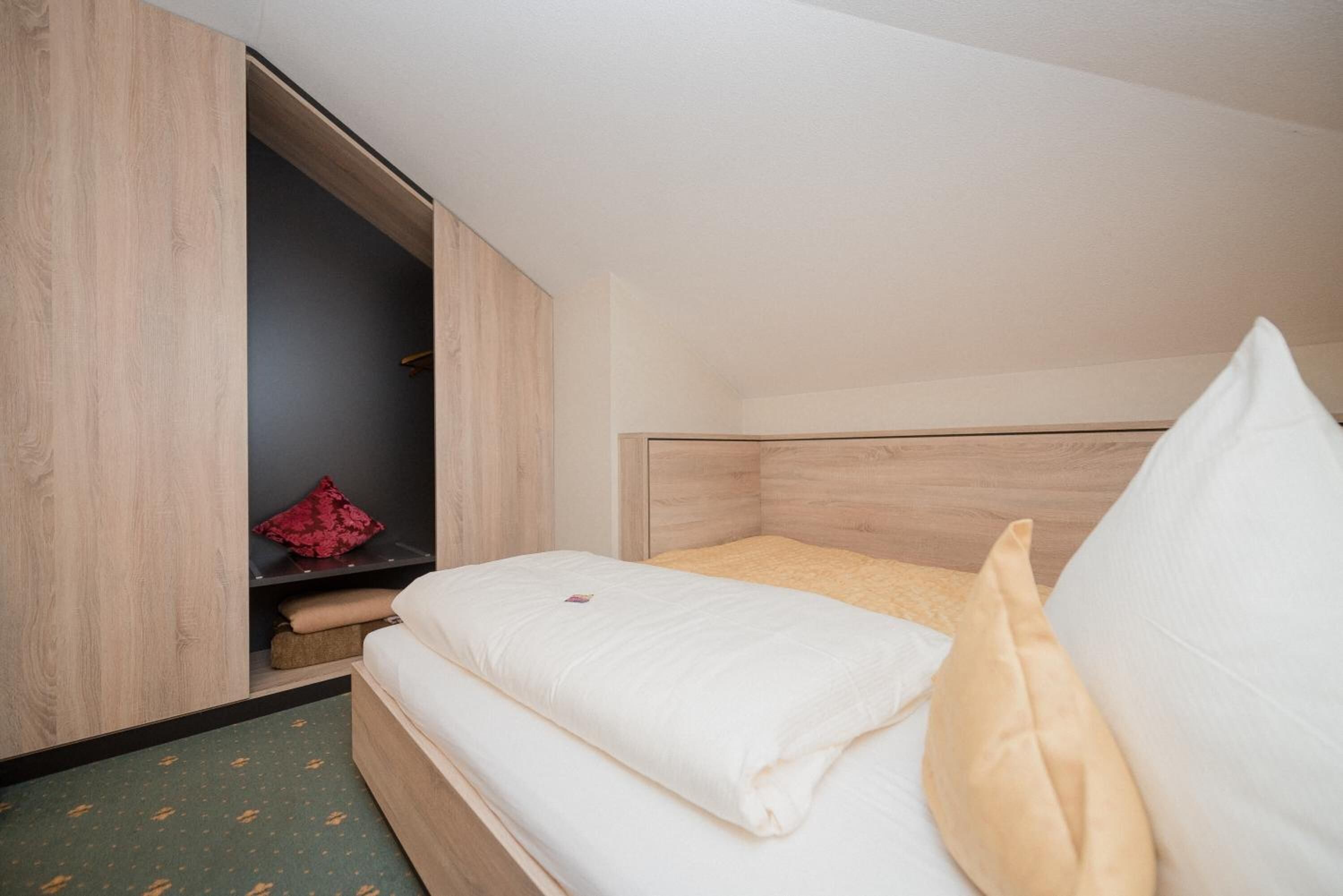 A small, neatly arranged bedroom with a wooden headboard, a bed with white bedding, and a red pillow on a shelf.