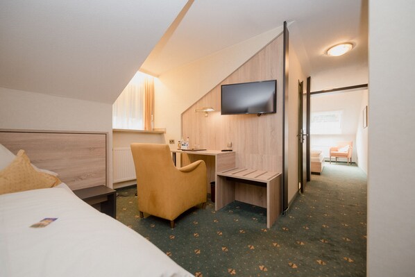 A hotel room with a bed, a desk, a chair, a TV, and a view of the hallway.