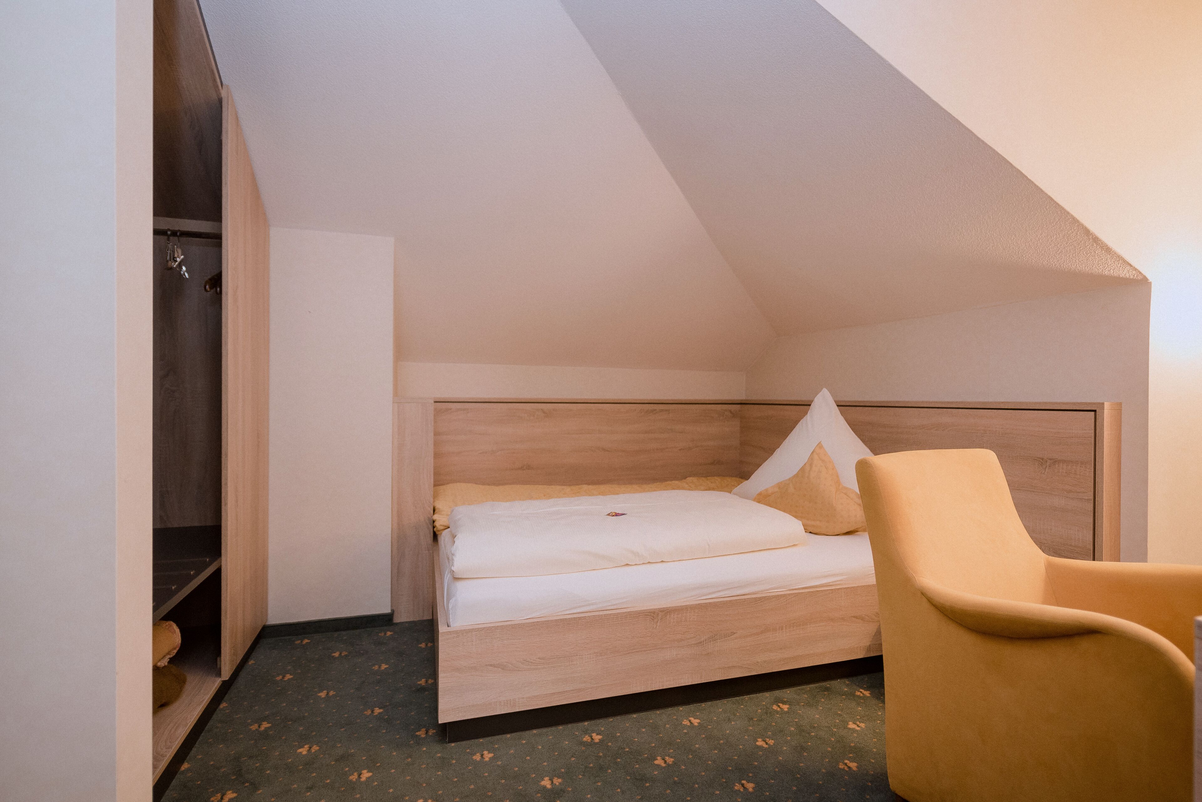 A small, single-bed room with a wooden headboard, a yellow chair, and a closet with hanging items.
