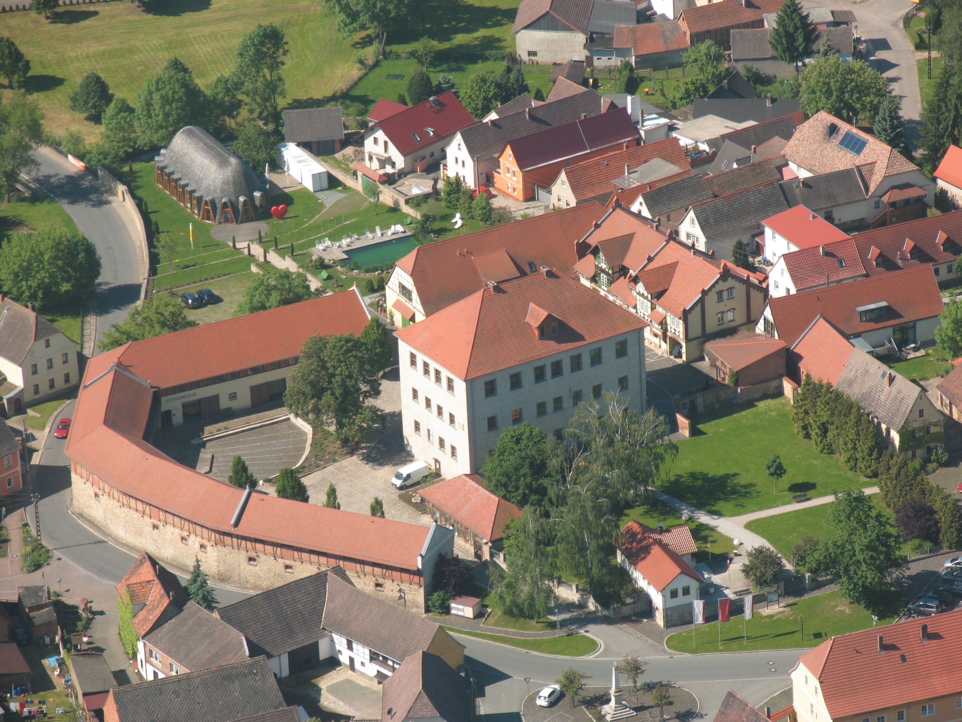 Une vue aérienne d’une ville avec un imposant bâtiment ressemblant à un château, doté d’un toit rouge, entouré d’immeubles résidentiels aux toits également rouges.