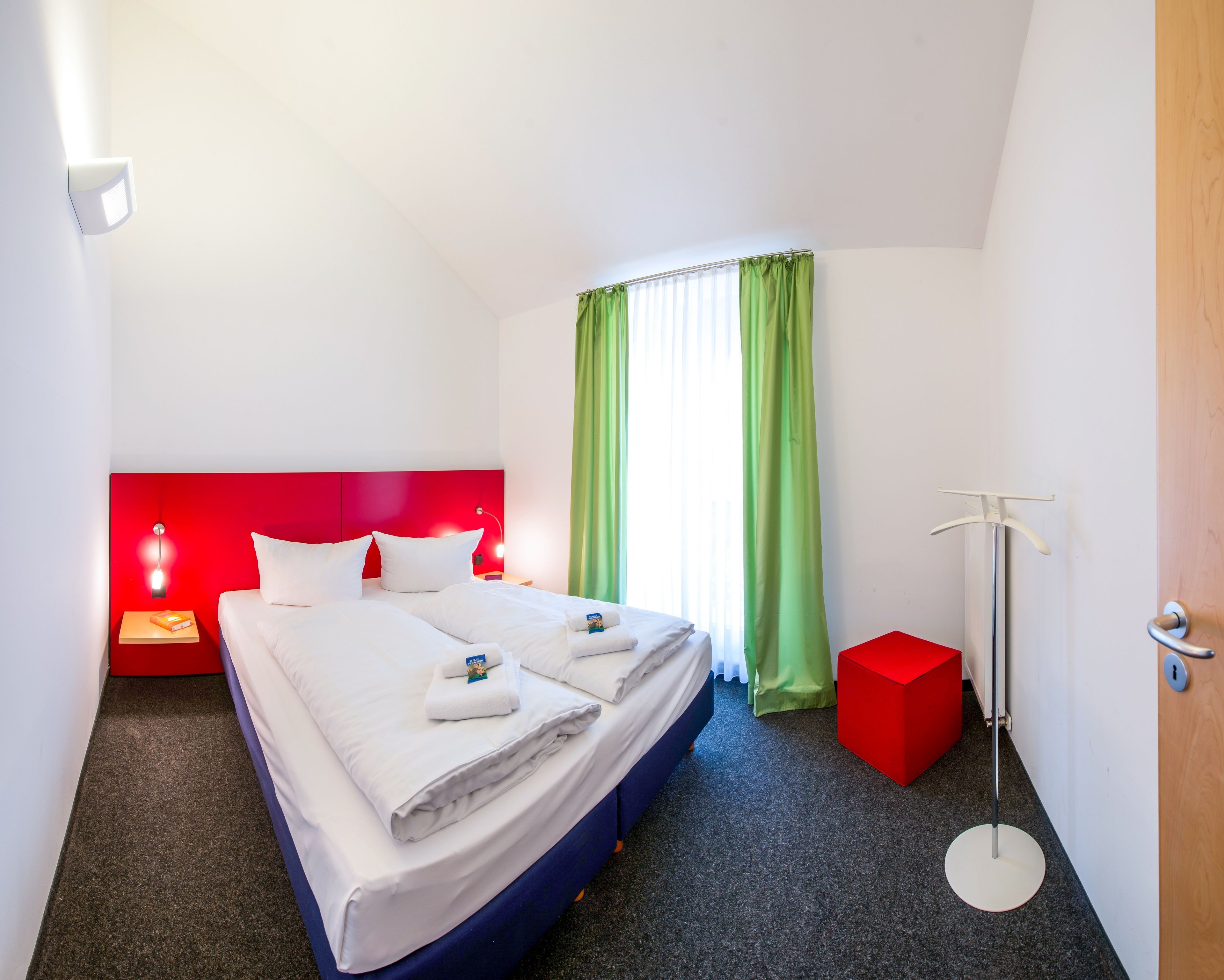 A hotel room with a bed, a red headboard, a nightstand with a lamp, a red stool, a floor lamp, and a window with green curtains.