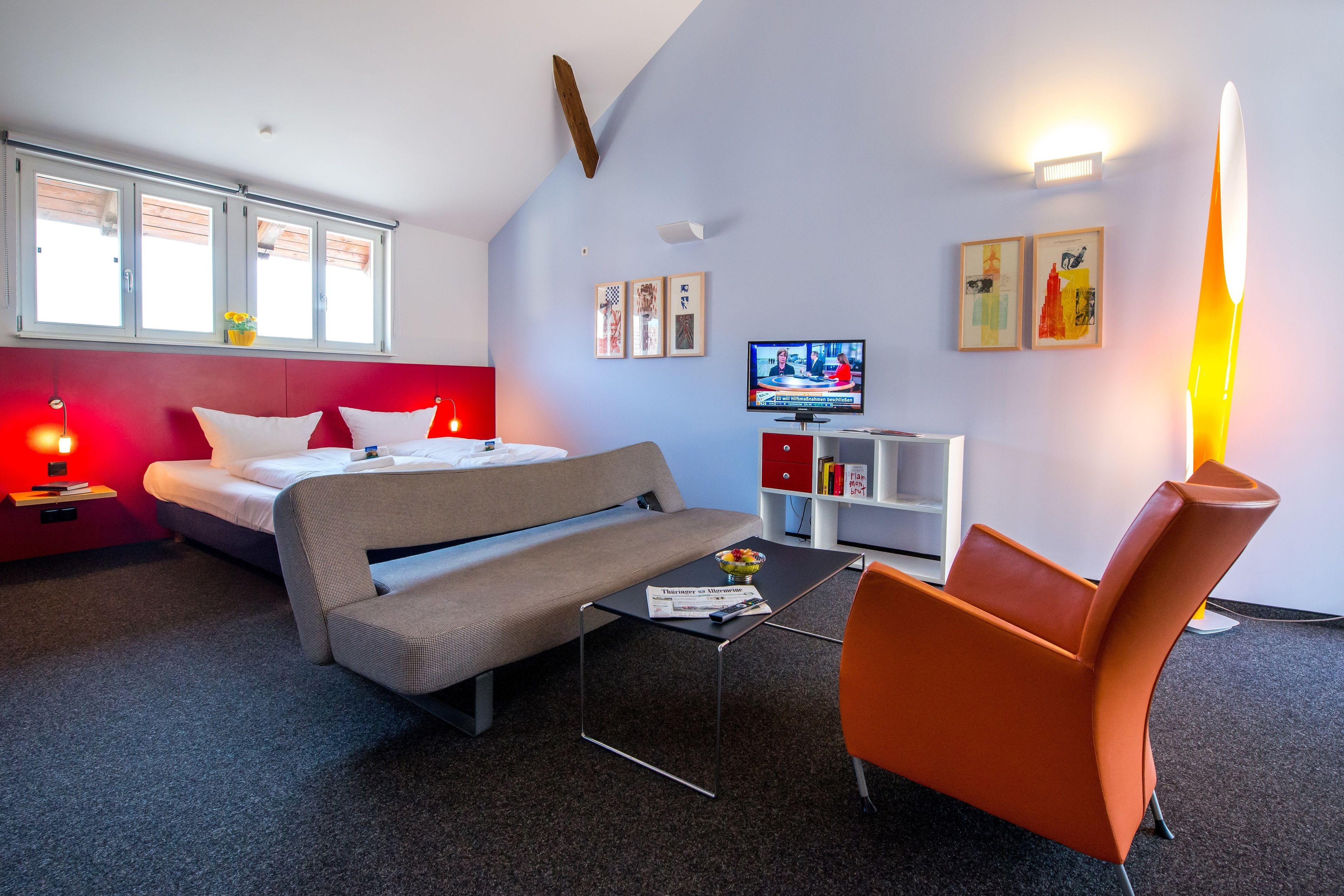 A modern hotel room with a bed, sofa, orange chair, and a small table with a bowl of fruit. There is a TV on the wall and a bookshelf with books.