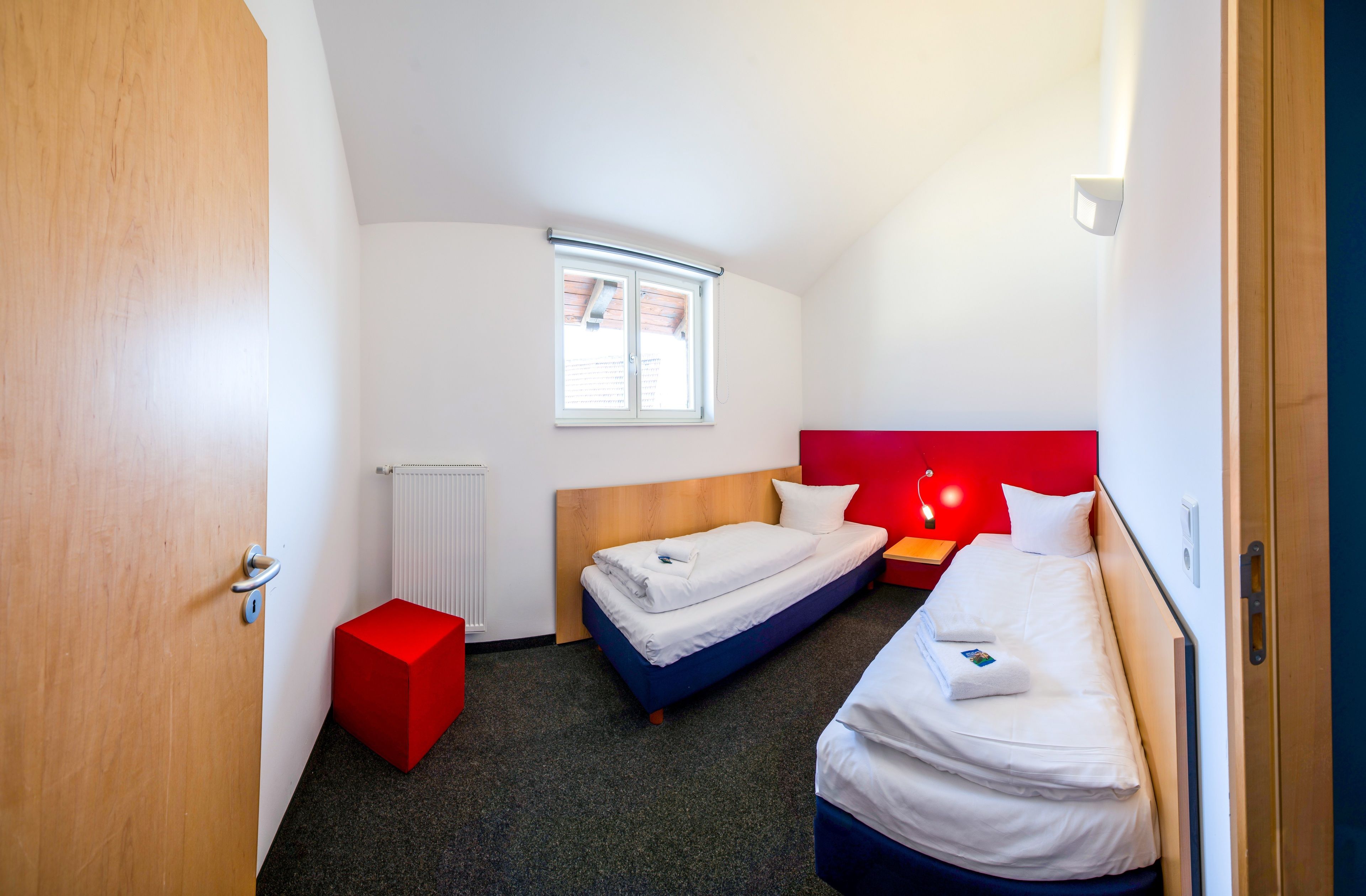 A compact hotel room with two beds, a red accent wall, a window, and a radiator.