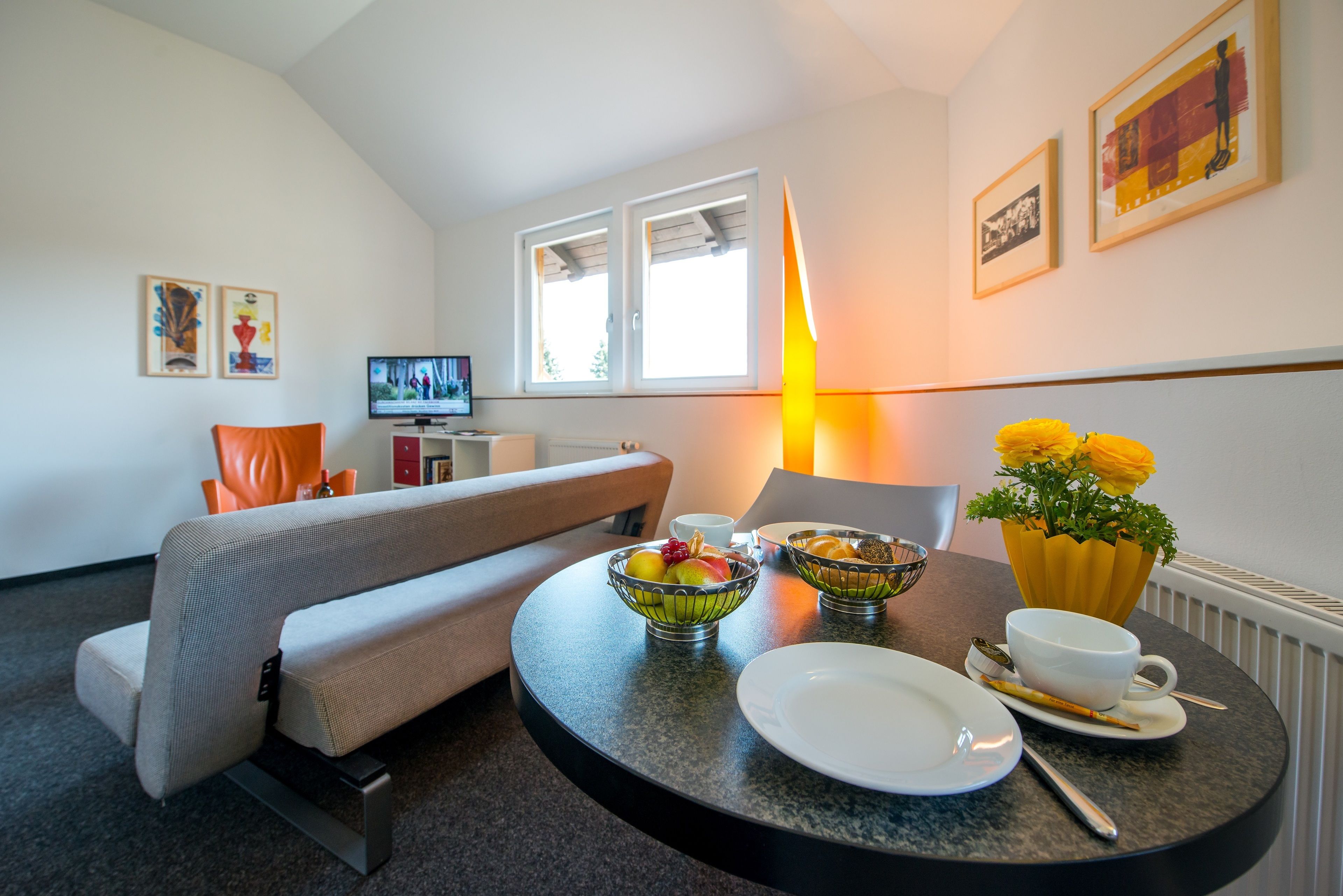 Une chambre d’hôtel moderne avec un canapé, une table à manger agrémentée de corbeilles de fruits, une tasse à café et un vase à fleurs.