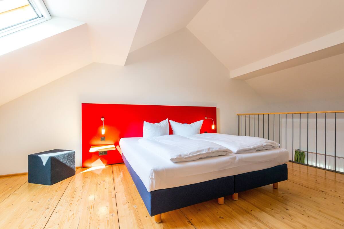 Een moderne slaapkamer met een schuine kap, een groot bed met witte lakens, een rode accentmuur en een dakraam.