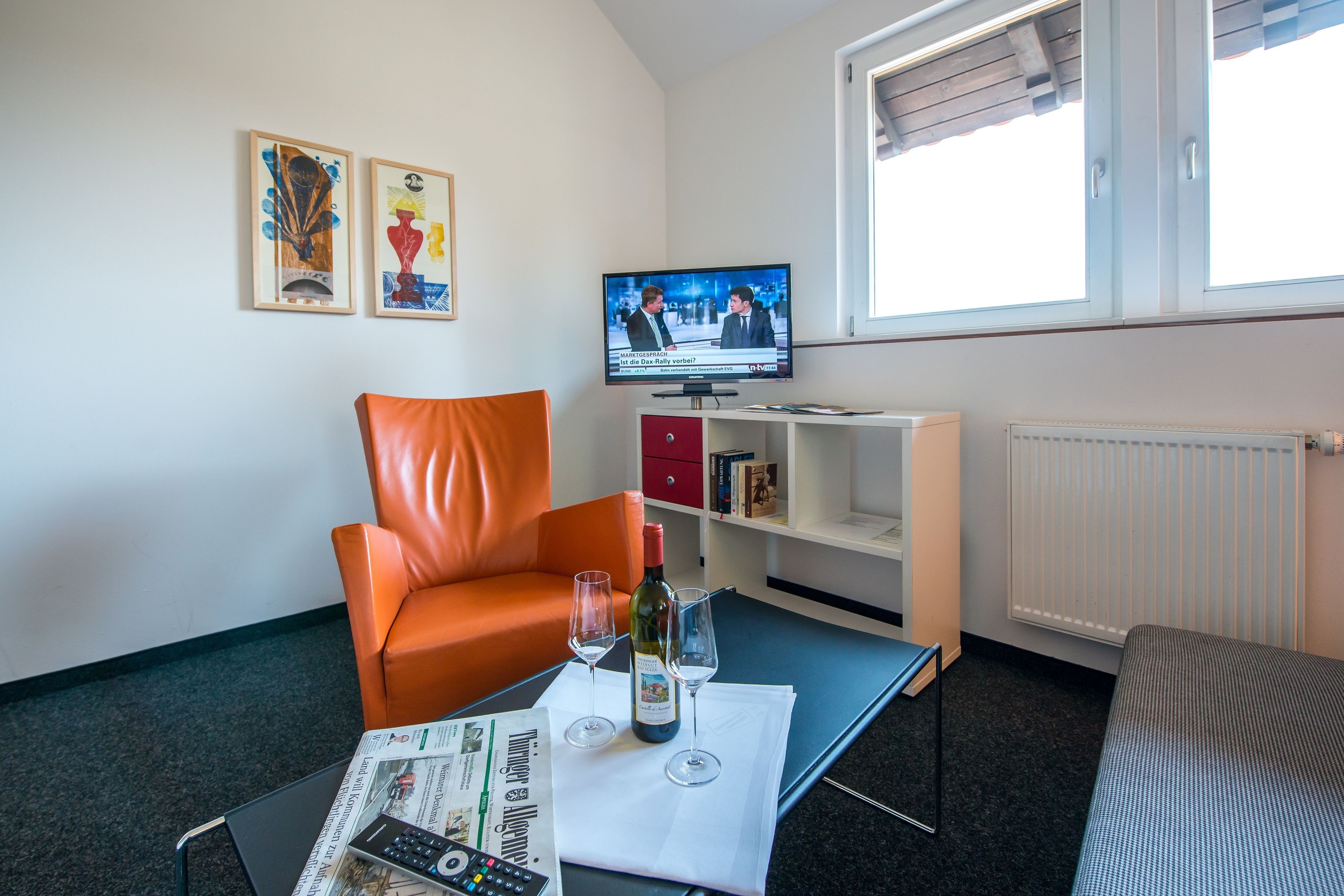 A modern living room with a leather armchair, a small table with a newspaper, wine glasses, and a TV displaying a news program.