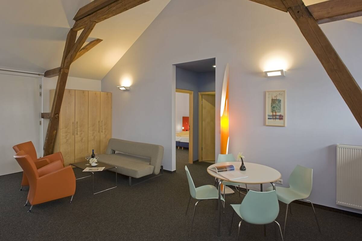 Een kamer met een hoge plafond, zichtbare houten balken, een bank, een tafel met stoelen en een lamp.