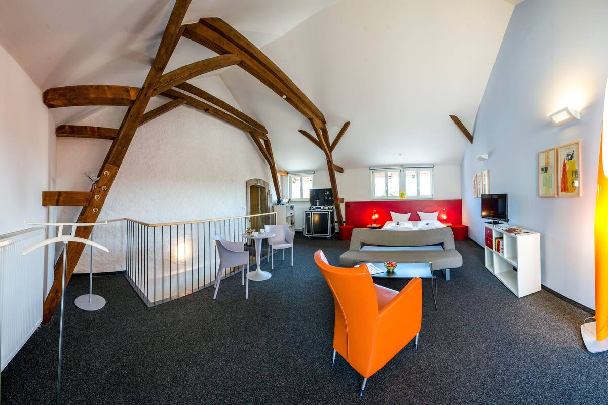 Een modern interieur met zichtbare houten balken, een bank, een feloranje stoel en een staande lamp.