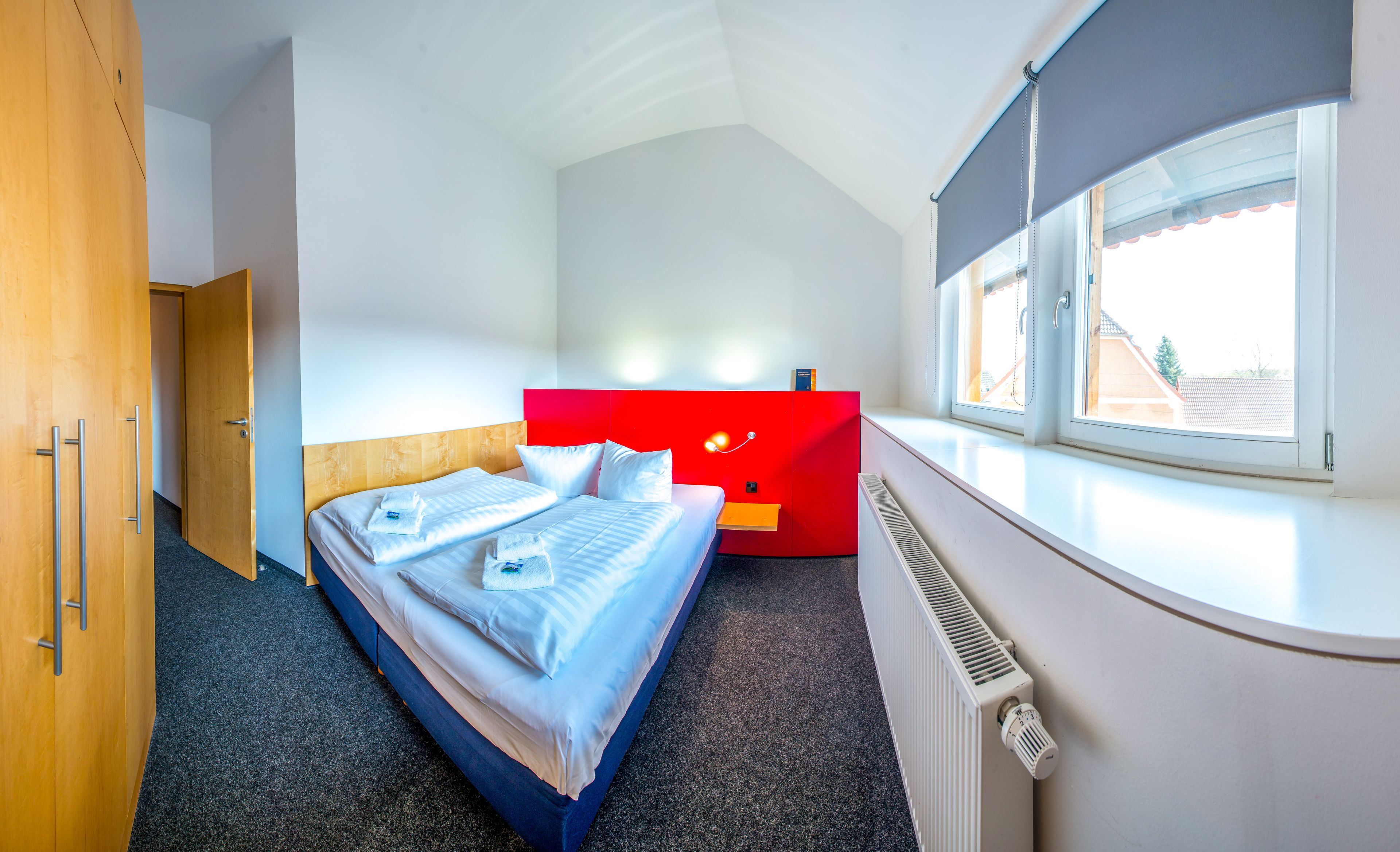 A modern hotel room with a bed, a red accent wall, a window with blinds, and a radiator.