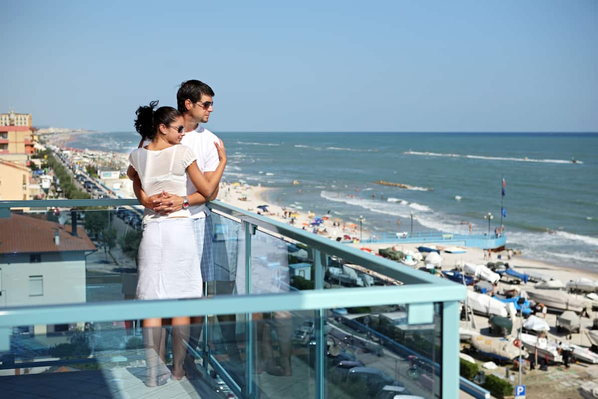 Un couple se tient sur un balcon donnant sur une plage, avec des bateaux et un ciel dégagé.