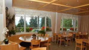 A dining area with a round table and chairs, large windows with curtains, and potted plants. - Hotel Alvier (Wartau)