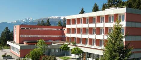 A multi-story building with a red and white facade, surrounded by greenery and a mountainous backdrop.