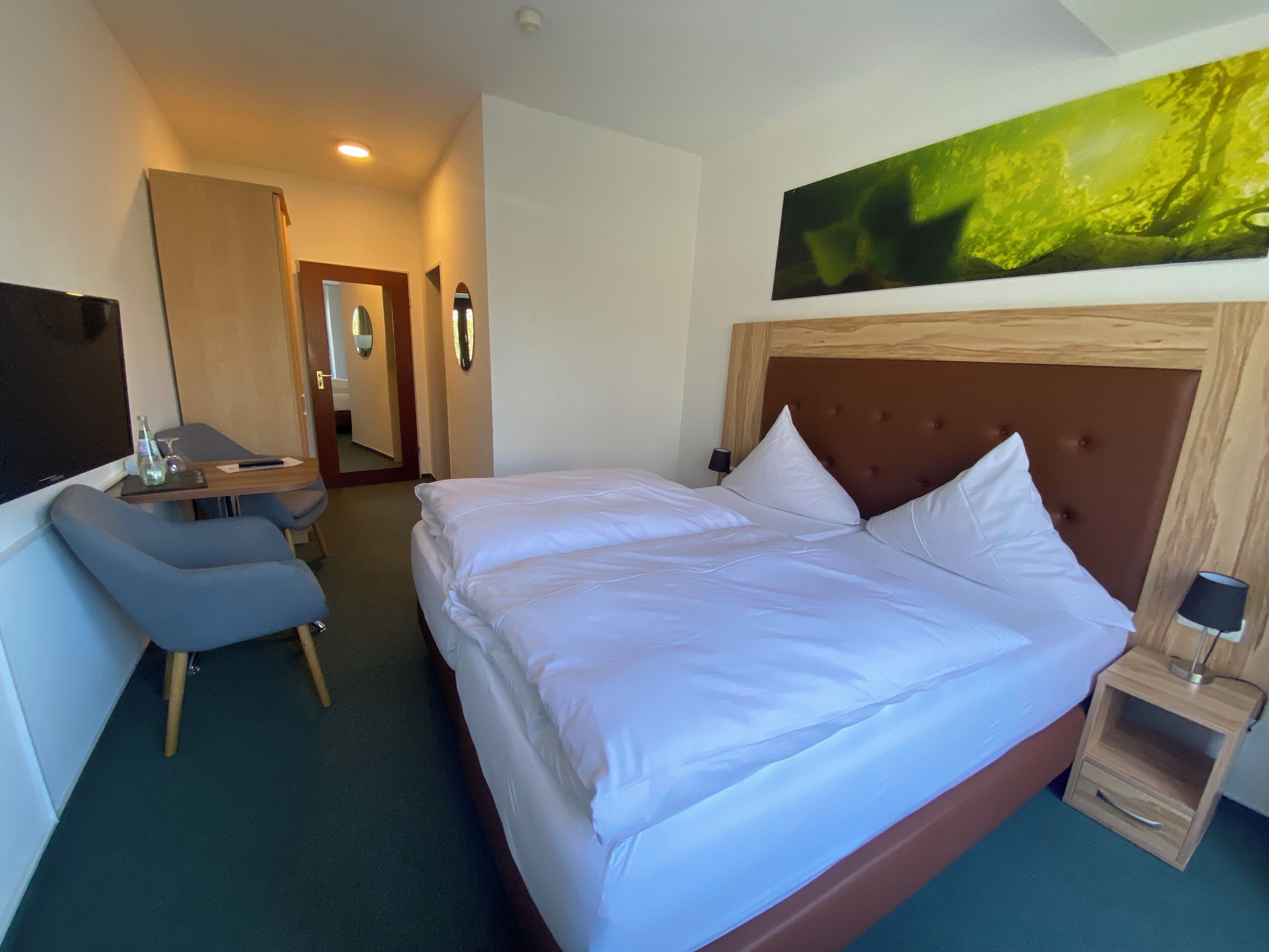 A hotel room with a bed, a desk, a chair, a nightstand, and a large painting on the wall.