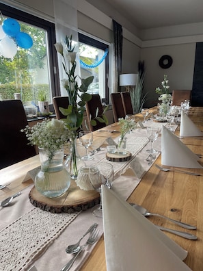 Uma mesa de jantar posta com copos, talheres e centros de mesa florais, com cadeiras ao redor.