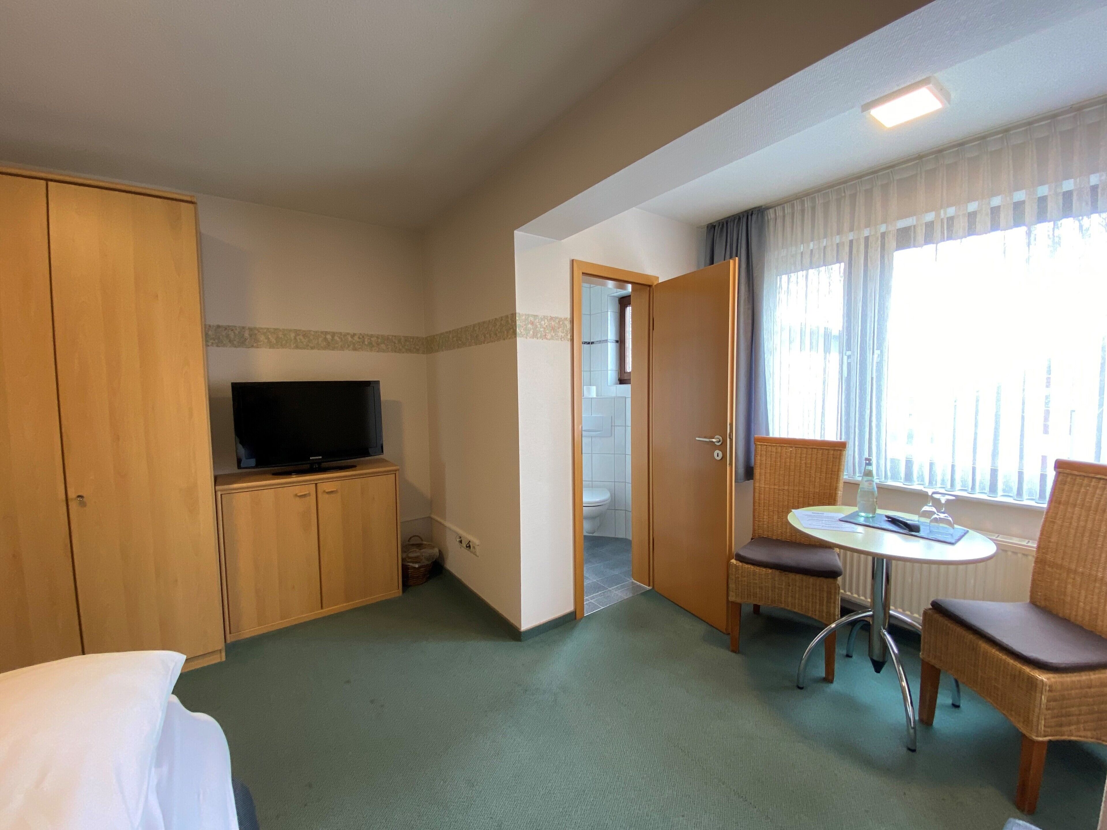 Una habitación de hotel con cama, televisión sobre un mueble de madera, una silla de mimbre, una mesita con una botella y una ventana con cortinas.