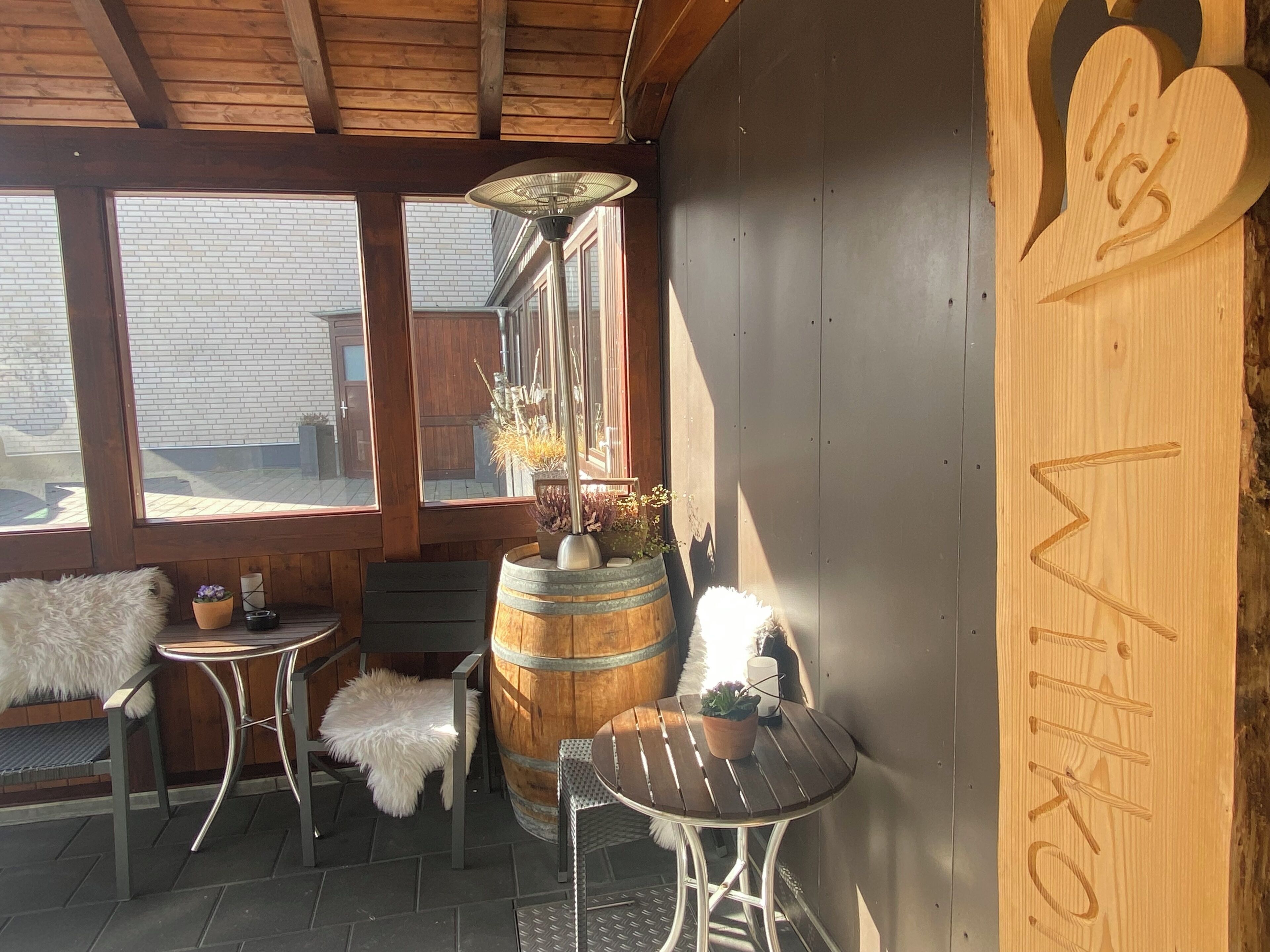 A covered outdoor seating area with a wooden ceiling, metal chairs, a small table, a decorative barrel, and a wall with carved text.