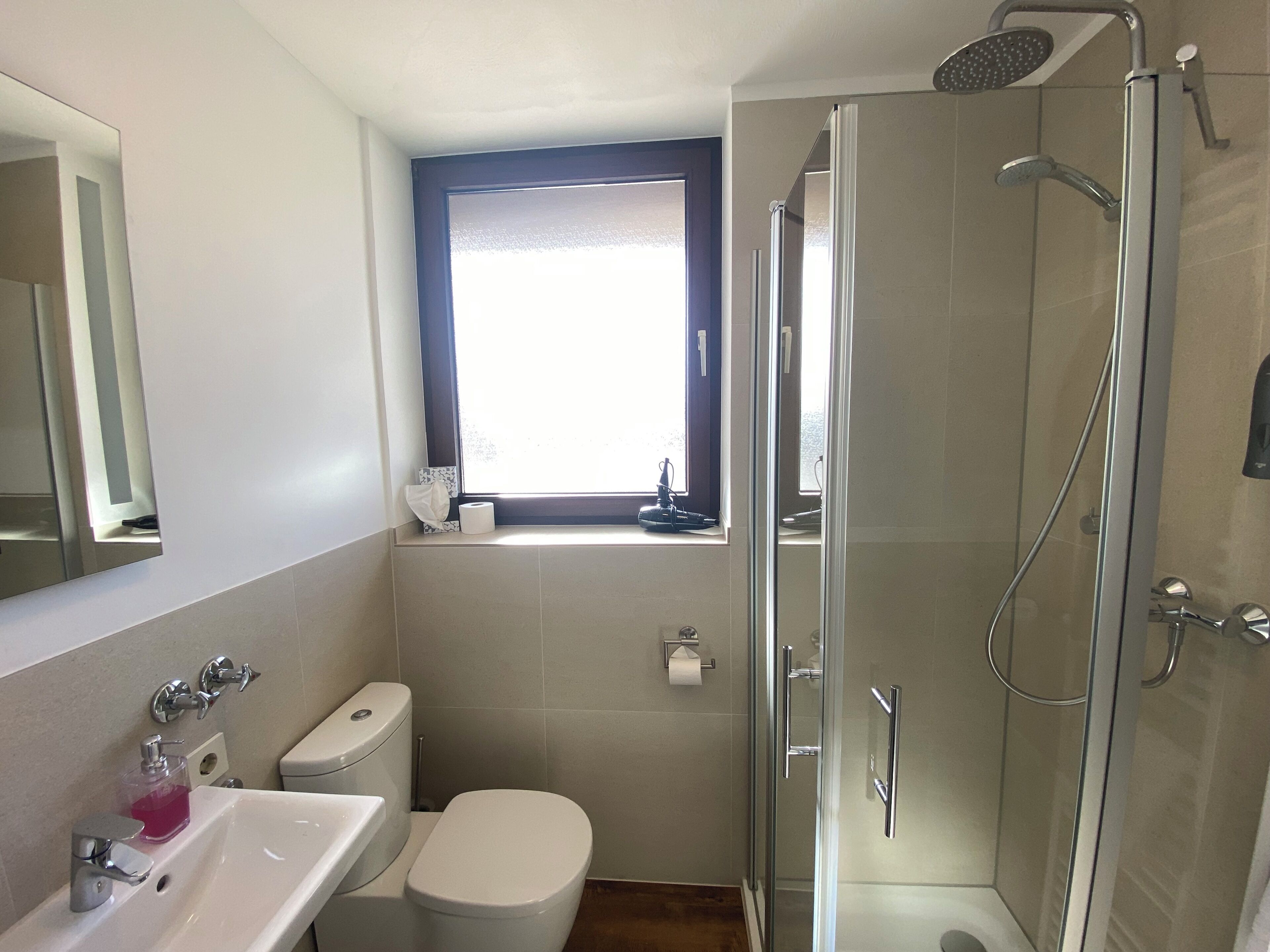 A bathroom with a toilet, sink, shower, and a window.
