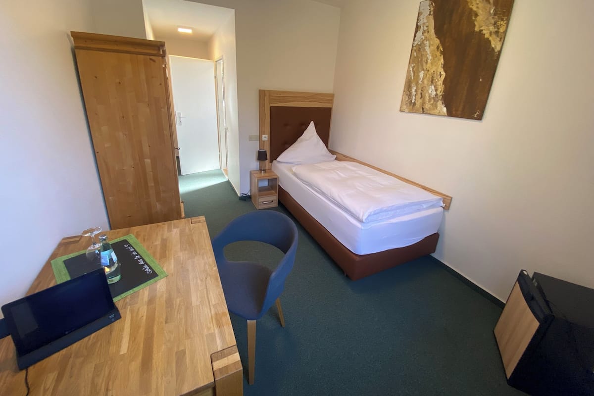 A hotel room with a single bed, a desk with a chair, a small table with a bottle, and a wall-mounted artwork.