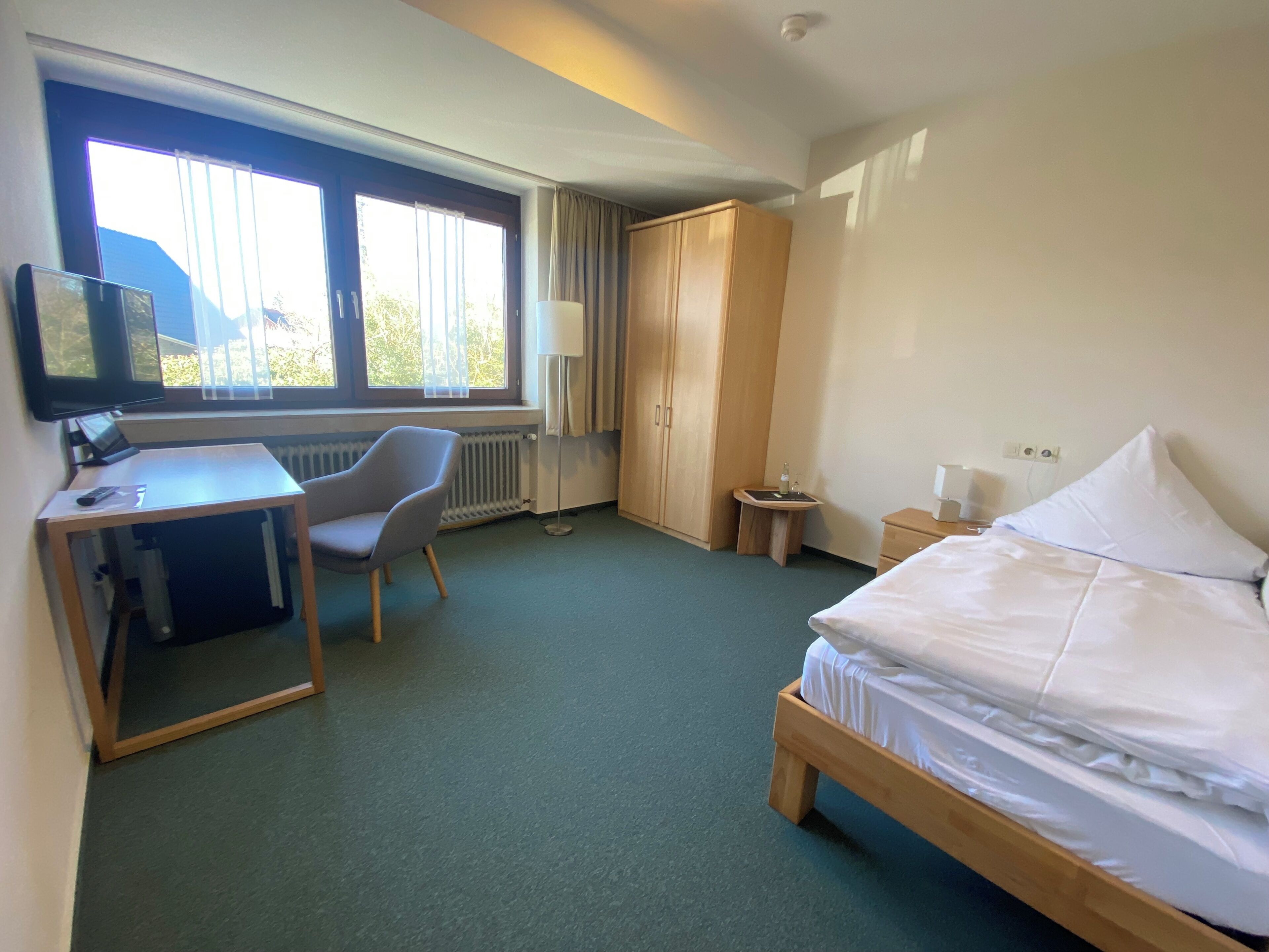 Habitación de hotel con cama, escritorio, silla, televisor, armario y una pequeña mesa con lámpara.