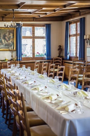 Comedor con una larga mesa dispuesta para una comida, sillas de madera y ventanas amplias con cortinas azules.