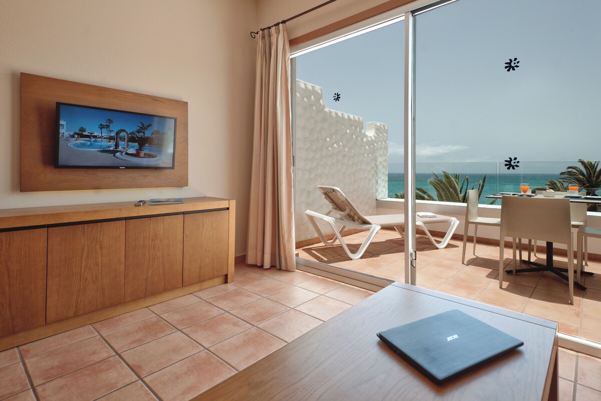Una habitación de hotel moderna con un televisor grande en la pared, un escritorio de madera, una computadora portátil, un balcón con vistas al mar y una zona de comedor con sillas y una mesa.