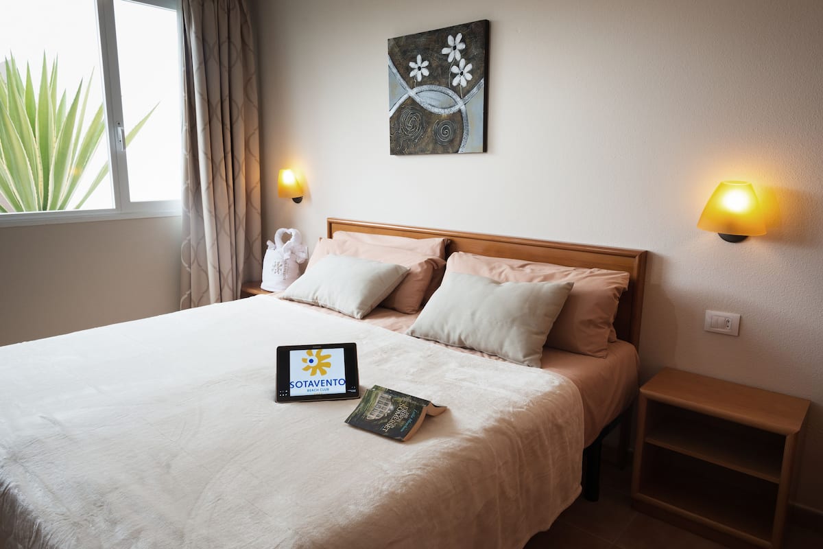 Una cama bien tendida con una tableta mostrando el logotipo de un club de playa, un libro y una lámpara de pared.
