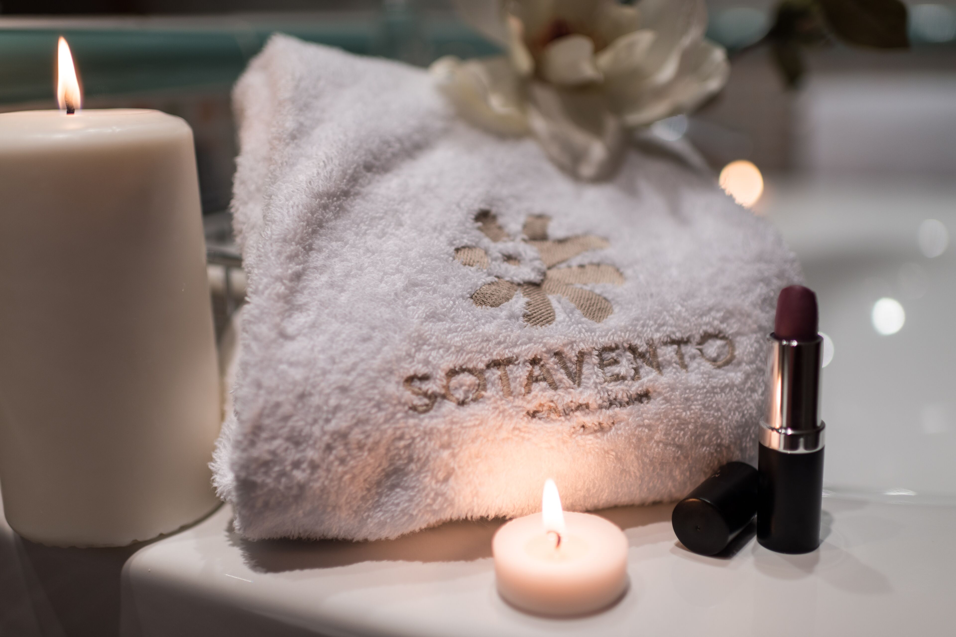 Una candela, un rossetto e un asciugamano con testo ricamato su una superficie bianca.