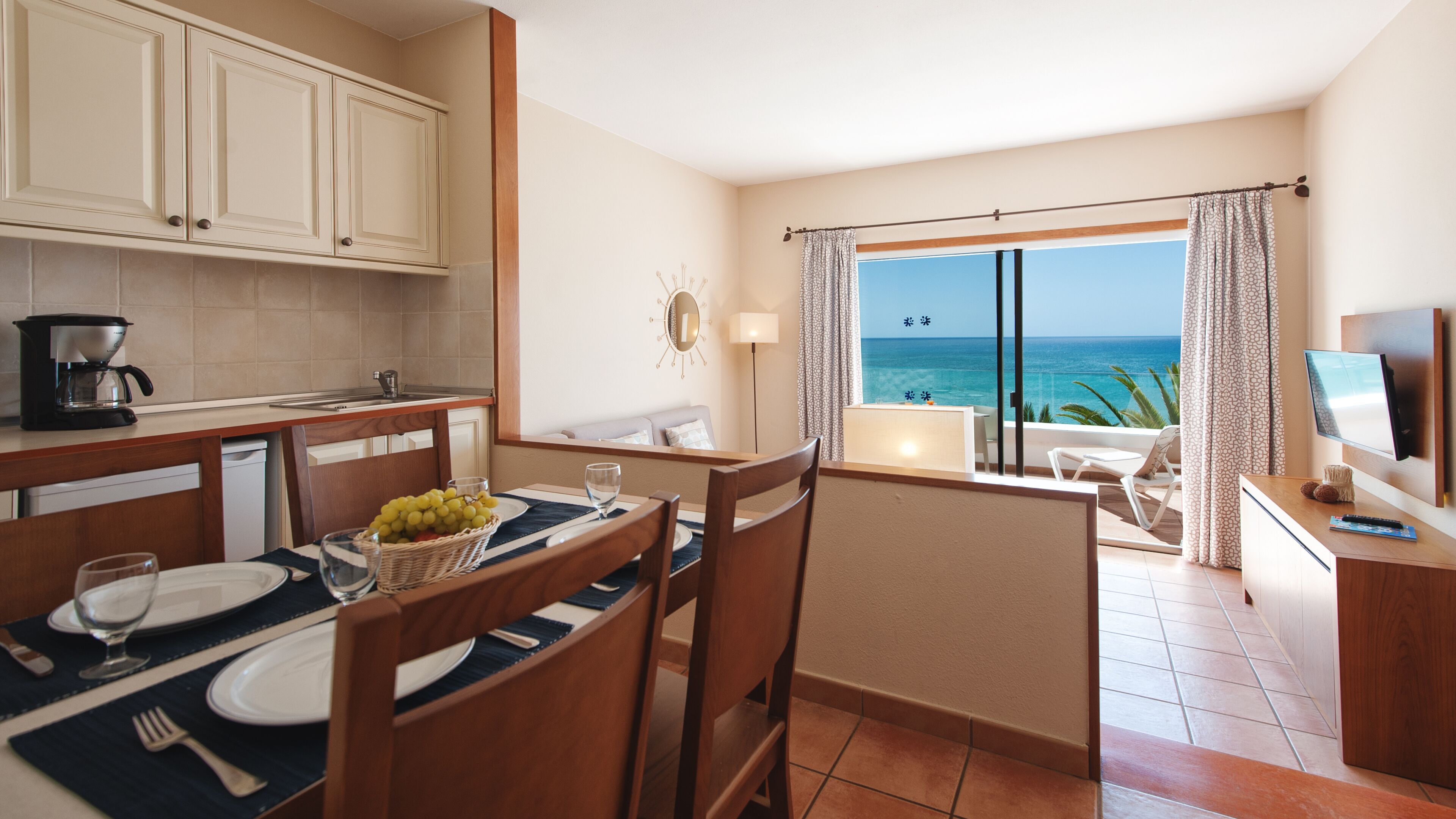A hotel room with a dining area, kitchenette, and a balcony with a view of the sea.