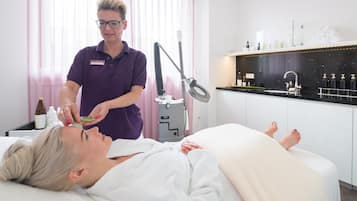 A person lying on a treatment bed receiving a facial treatment from a professional in a clinical setting with a modern, clean design.