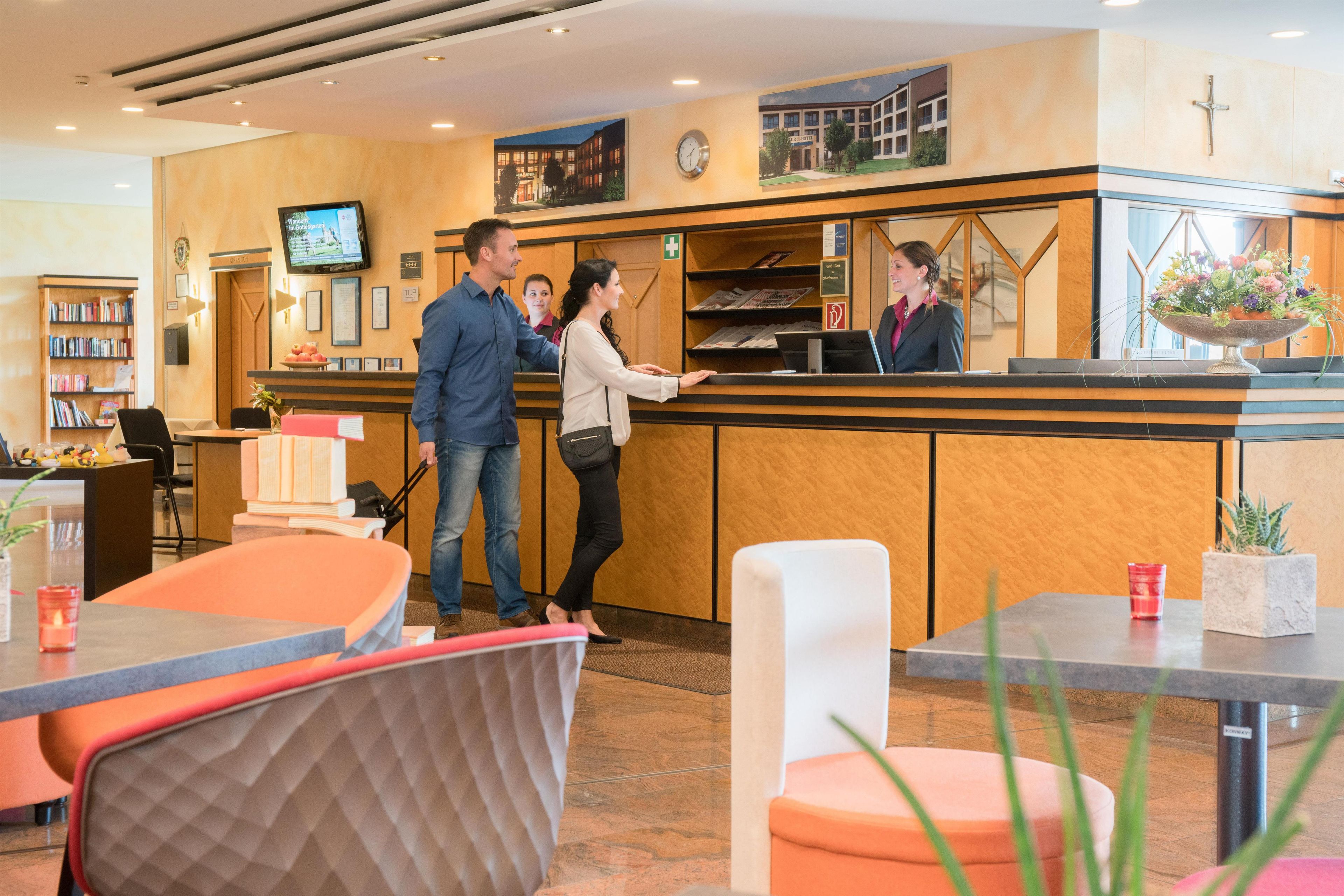 A hotel reception area with a reception desk, a television, a clock, and a cross on the wall.