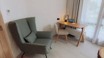 A modern room with a green armchair, a wooden desk with a lamp, and a view of the outdoors through sheer curtains.