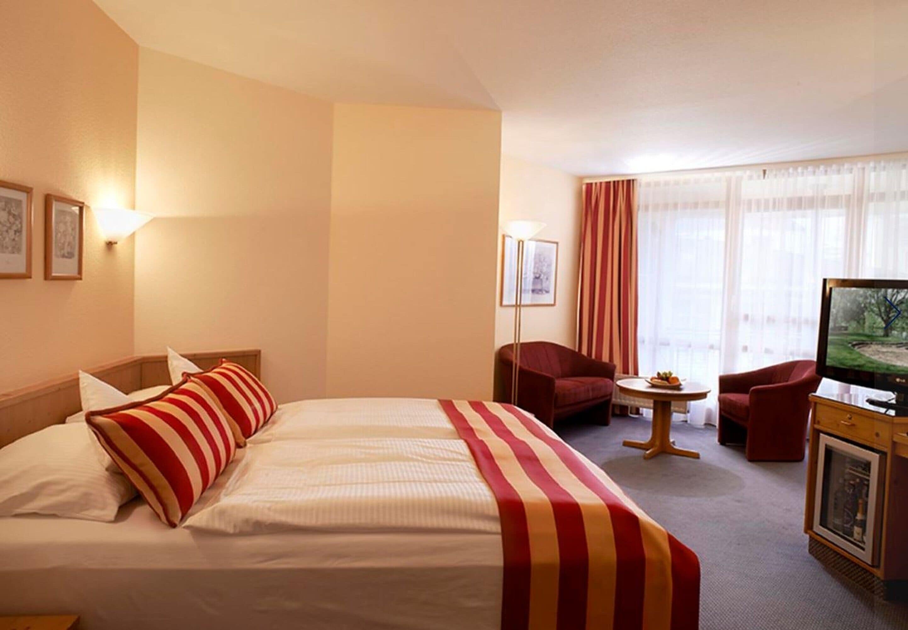 Ett hotellrum med en stor säng, två röd- och vitrandiga kuddar, en tv, ett litet bord med en fruktskål och två röda fåtöljer.