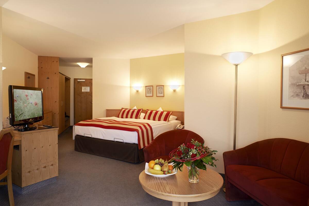 A hotel room with a bed, a desk with a computer, a round table with a fruit bowl and flowers, a red sofa, and a lamp.