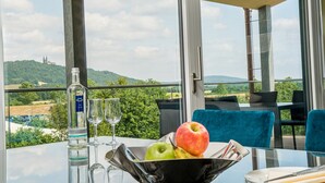 Une table avec une bouteille, des verres et un saladier de fruits, placée sur un balcon d’où l’on aperçoit une colline et des arbres au loin.