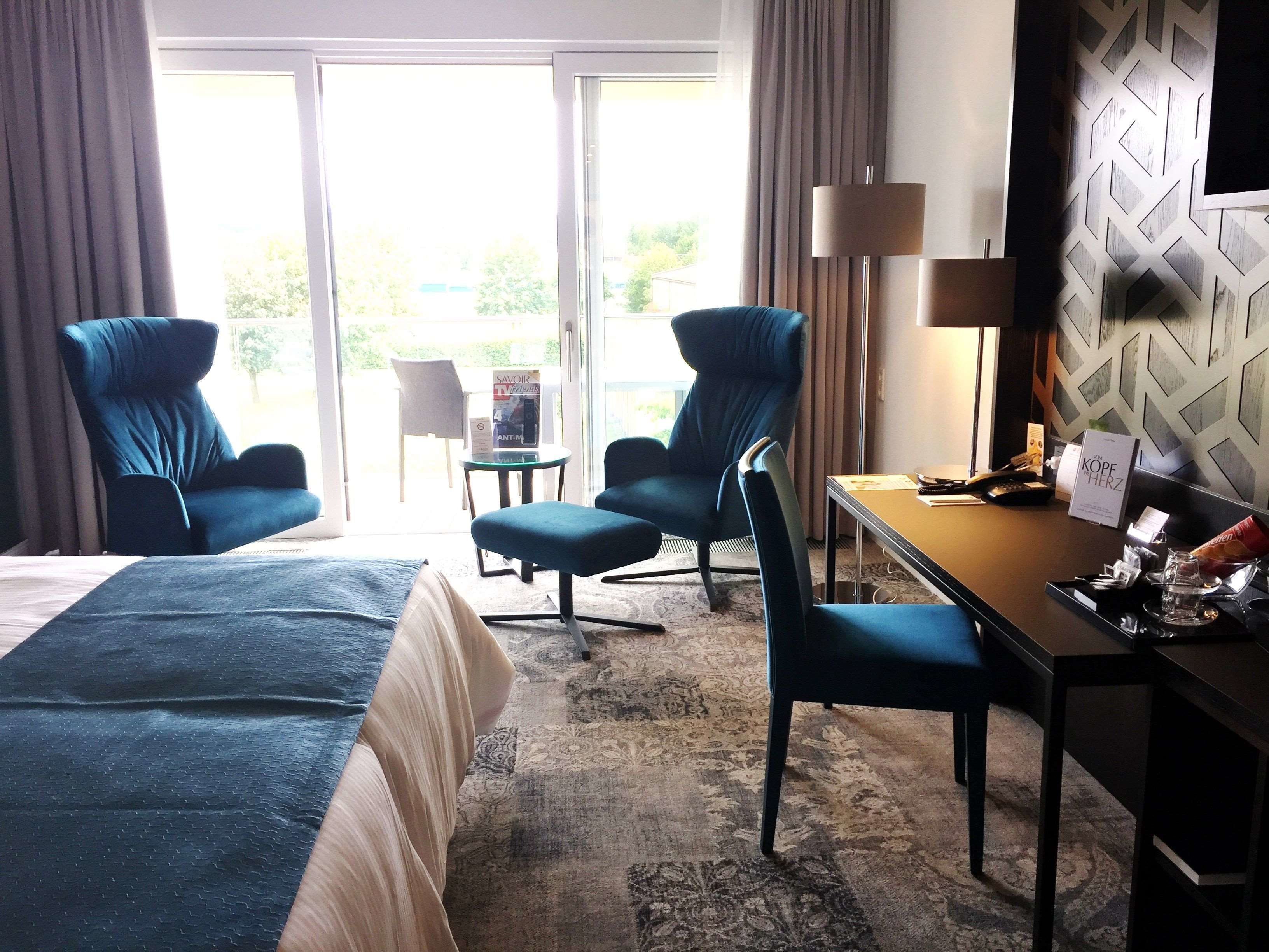 Ein Hotelzimmer mit einem Bett, zwei Sesseln, einem kleinen Tisch, einem Schreibtisch, einer Lampe und Blick ins Freie.