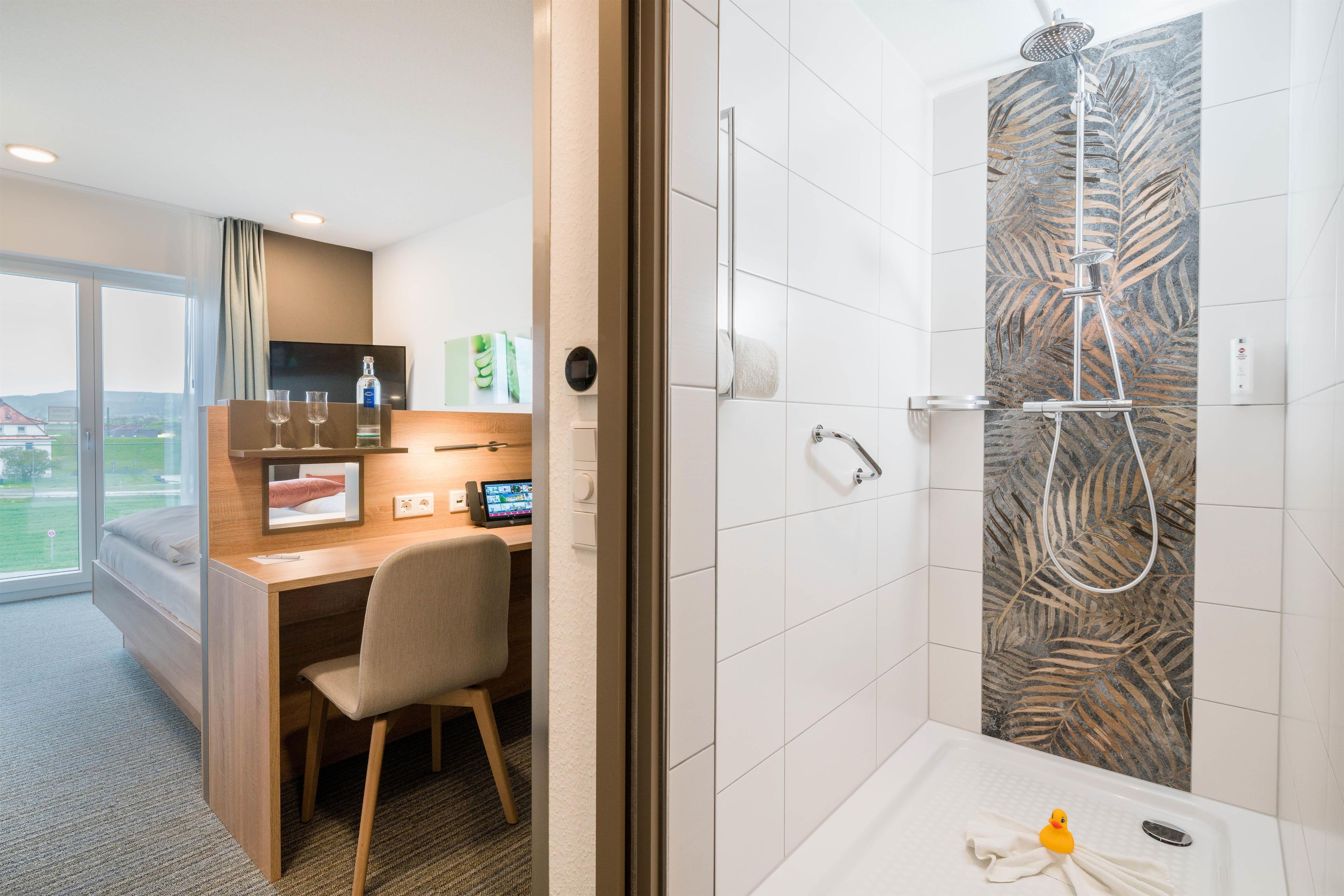A hotel room with a desk, chair, and a shower area with a leaf-patterned wall.