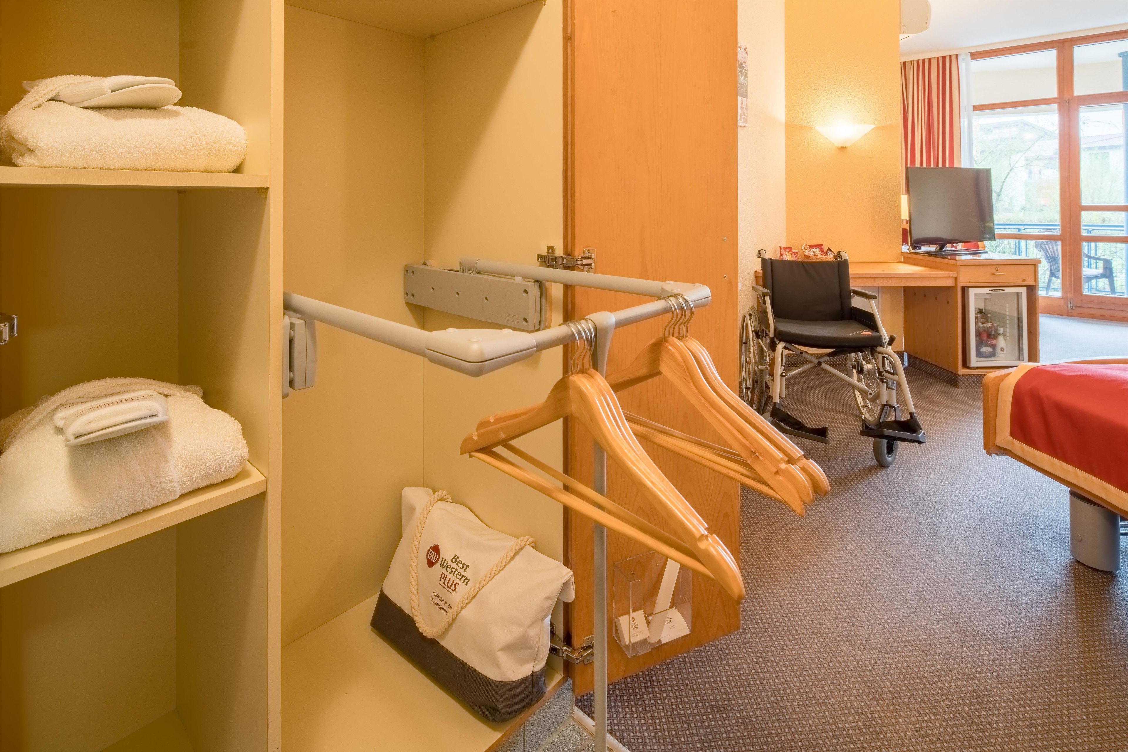A hotel room with a wheelchair, a desk with a computer, a bed, and a shelf with towels.