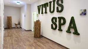 Un couloir doté d’un plancher en bois, un mur orné de l’inscription « VITUS SPA » réalisée à l’aide de motifs végétaux verts, et une porte au fond.
