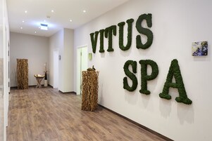 Un pasillo con piso de madera, una pared con 'VITUS SPA' escrito en follaje verde, y una puerta al final.
