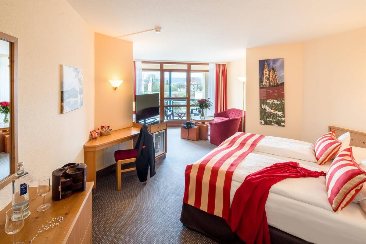 A hotel room with a bed, a desk with a chair, a TV, a window with a view, and a poster on the wall.